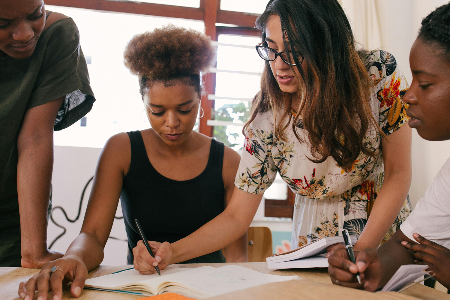 A close-up of a collaborative meeting where four women are engaged in reviewing notes. One woman is writing, while another is pointing at her notebook, offering guidance. They appear focused and engaged in their teamwork.
