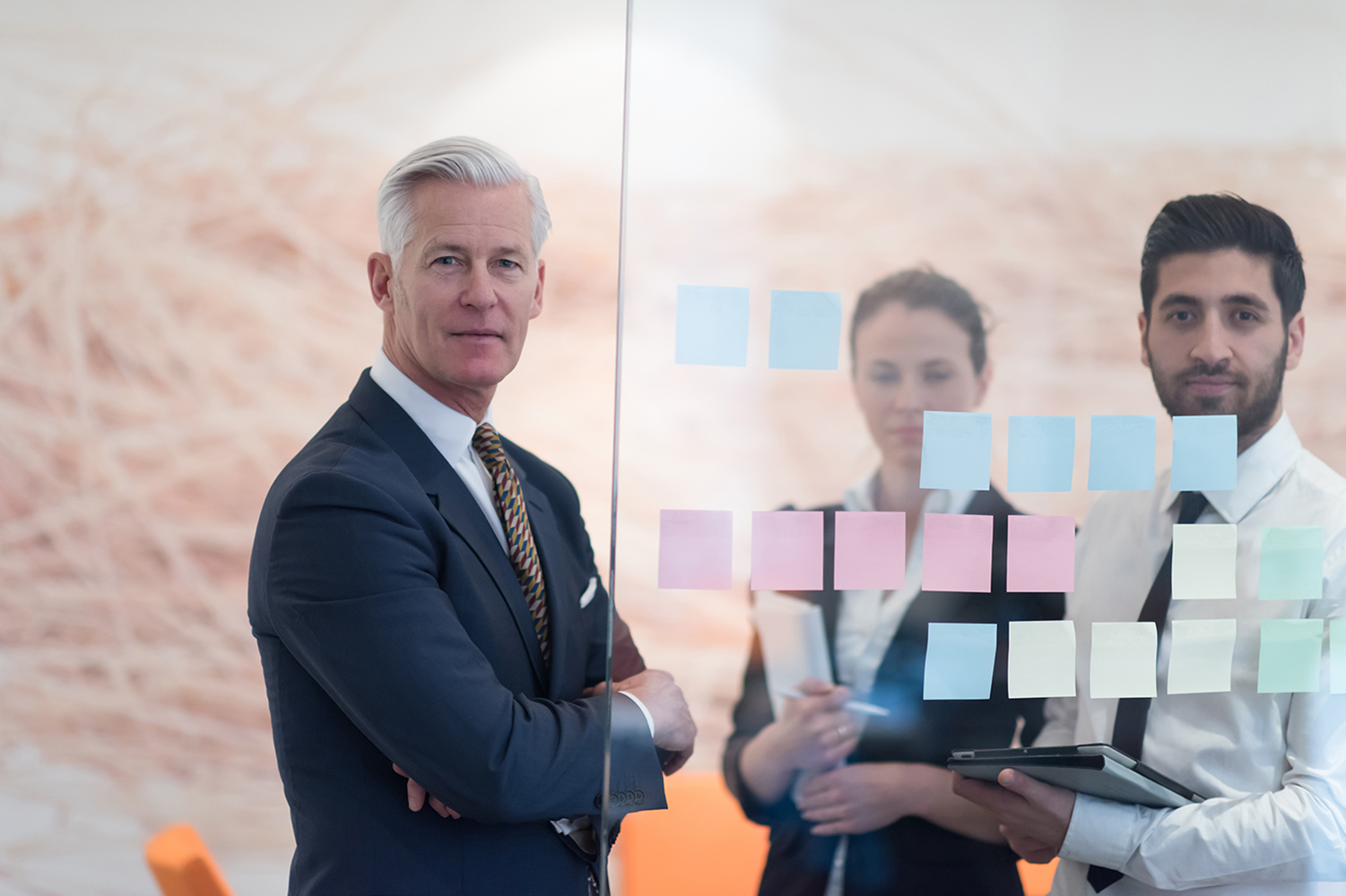 Senior executive standing in front of a glass wall covered with colorful sticky notes, while two younger professionals stand behind, actively engaged in a planning or brainstorming session.