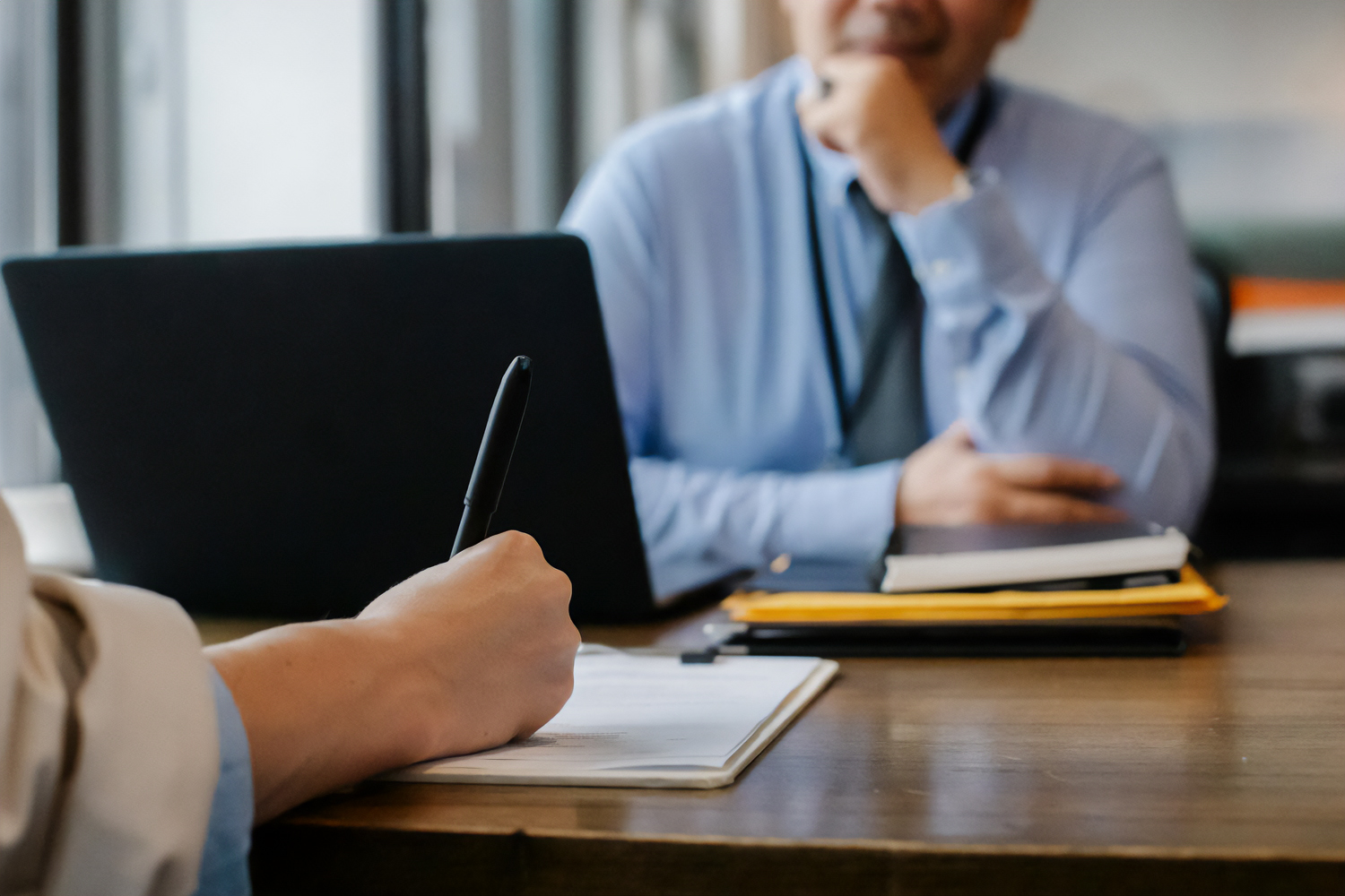 Close-up of a person taking notes during a professional interview, with a laptop and documents on the table, representing a background check or hiring process.
