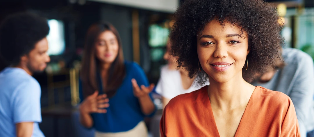 Confident young professional in an orange blouse standing in focus while colleagues discuss in the background, highlighting workplace integrity and leadership assessments.
