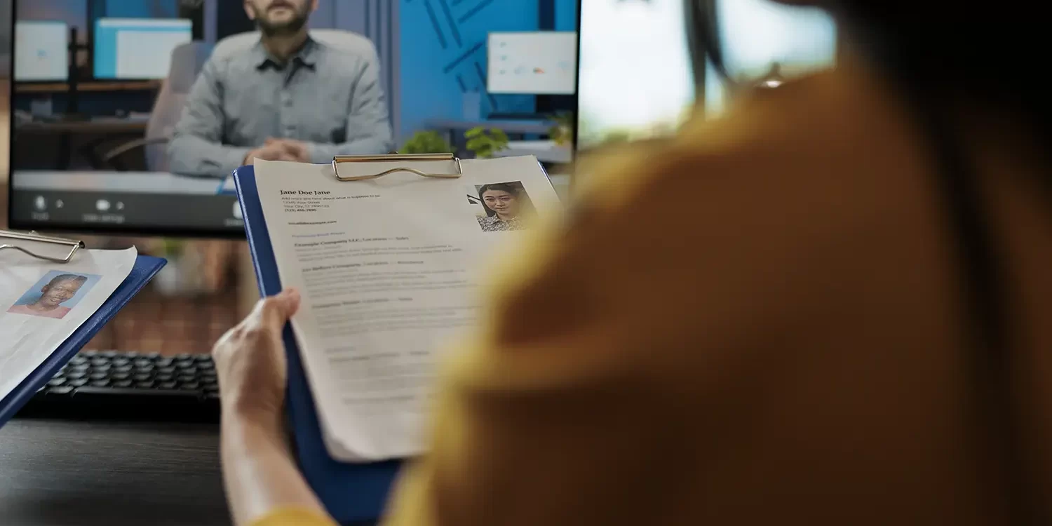 Recruiter conducting virtual interview while reviewing printed resumes, using hiring platform for candidate evaluation and selection.