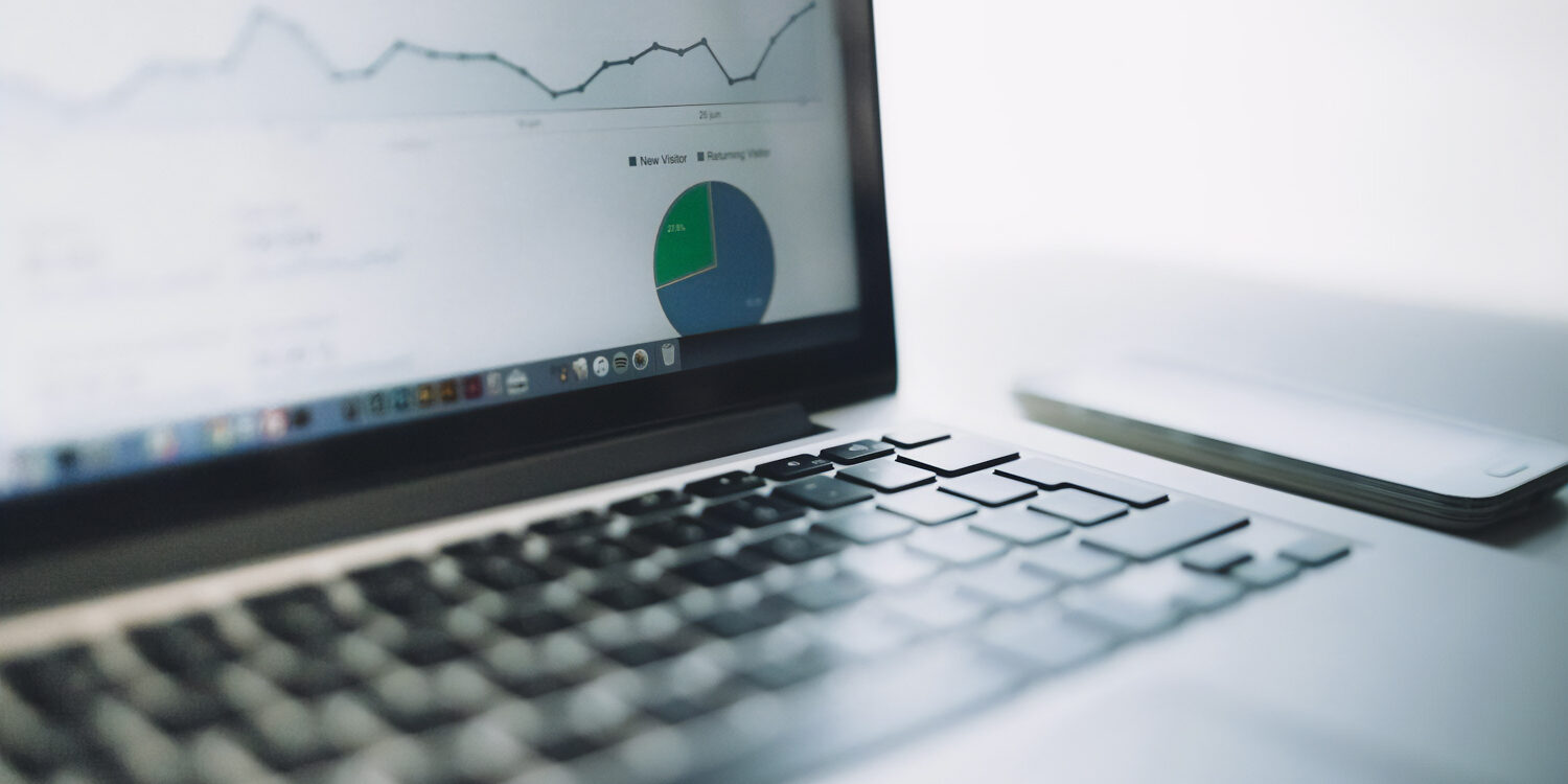 A close-up shot of a laptop displaying an analytics dashboard with a line chart and a pie chart, accompanied by a smartphone lying next to it on a clean white desk.