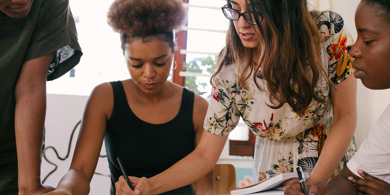 A close-up of a collaborative meeting where four women are engaged in reviewing notes. One woman is writing, while another is pointing at her notebook, offering guidance. They appear focused and engaged in their teamwork.