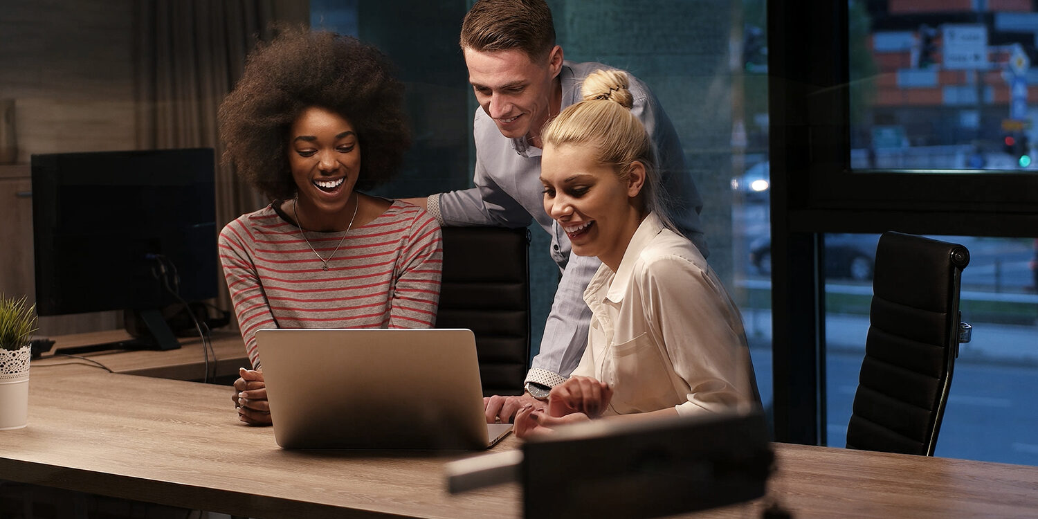 Three colleagues gathered around a laptop, smiling and collaborating in a modern office during the evening.