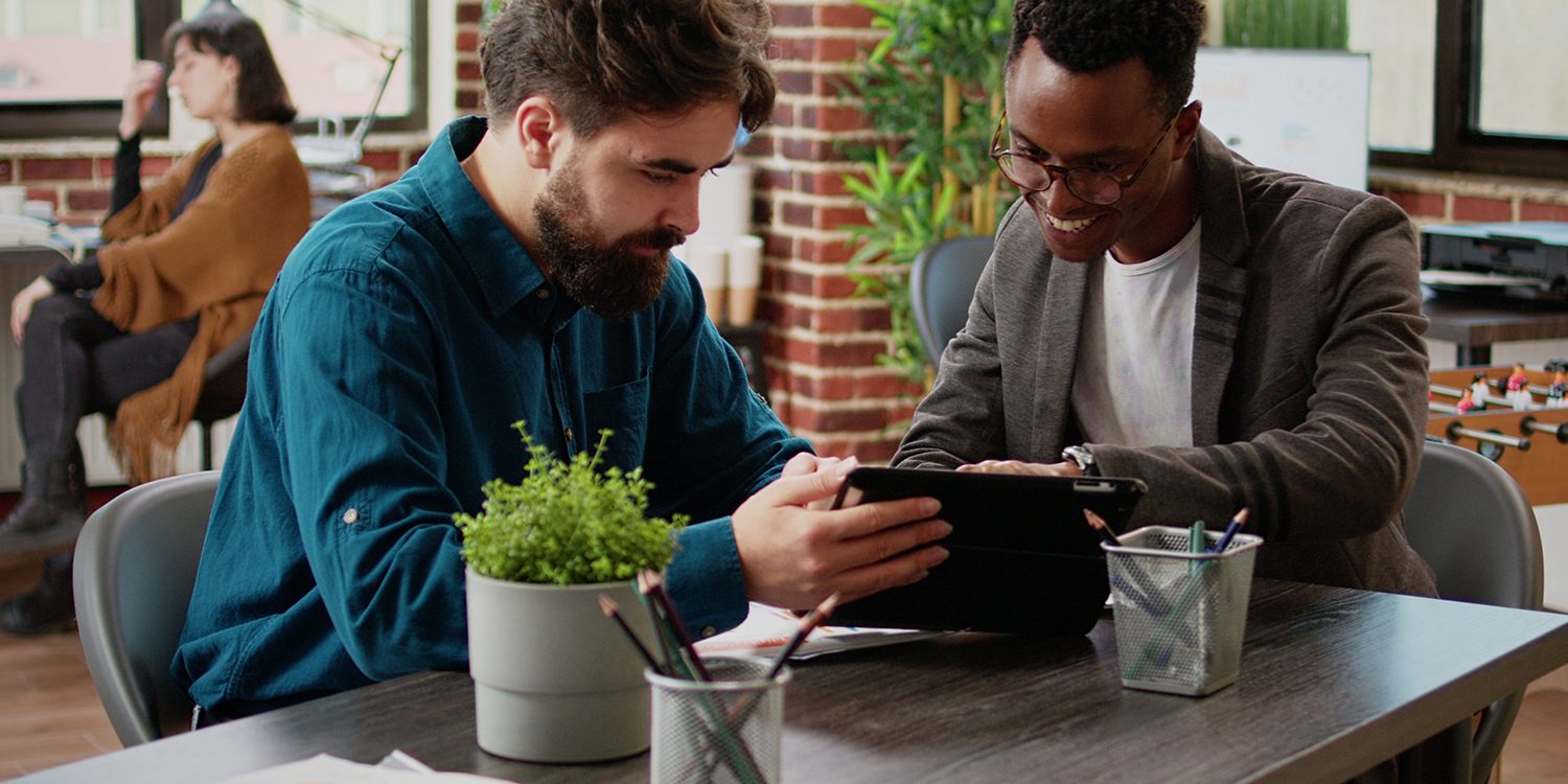 Two colleagues smiling and collaborating on a digital tablet at a modern office desk with paperwork and plants, while another person works in the background.