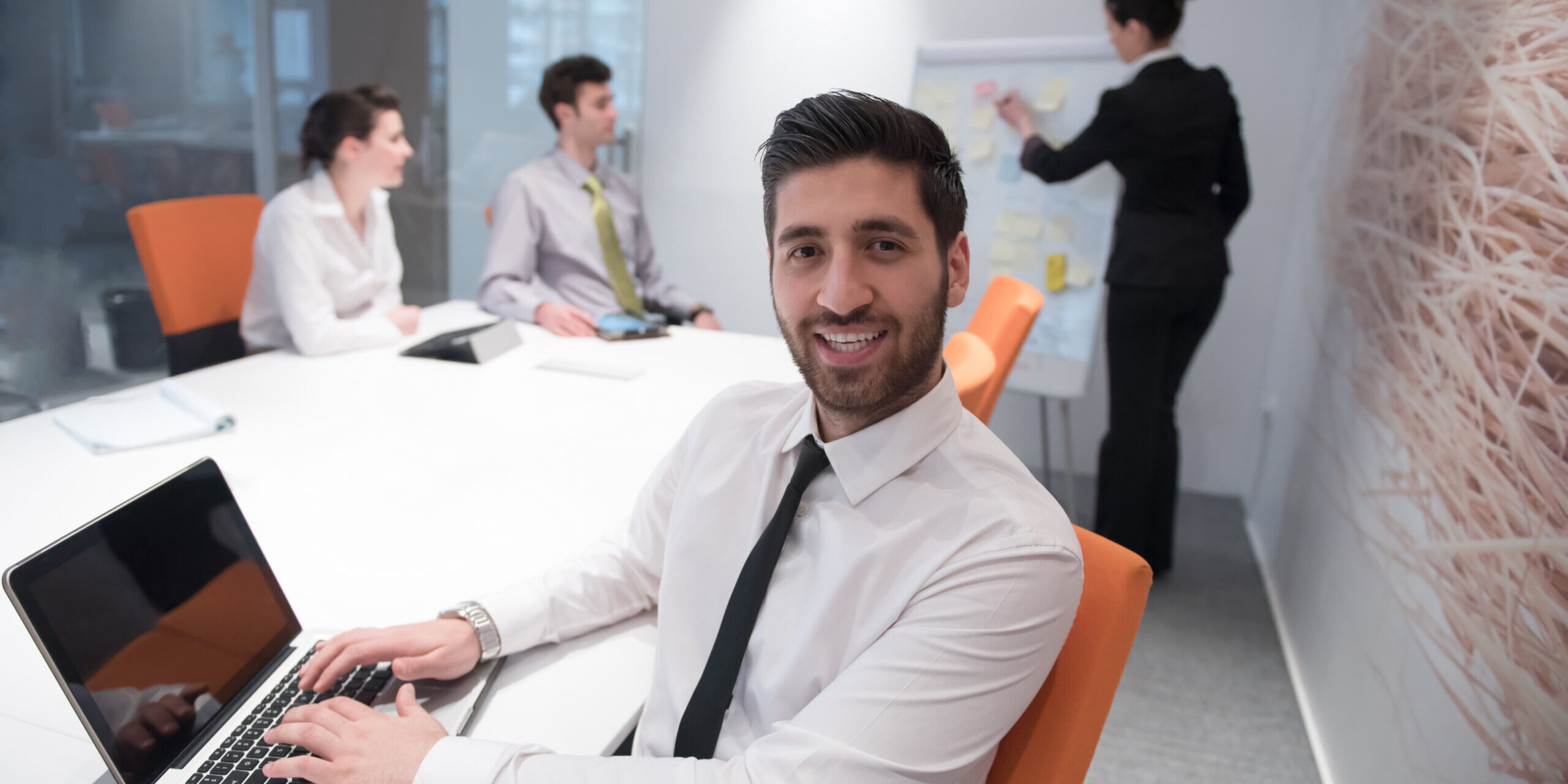 Confident young businessman in a white shirt and black tie smiling at the camera while typing on a laptop during a team meeting in a modern office conference room.
