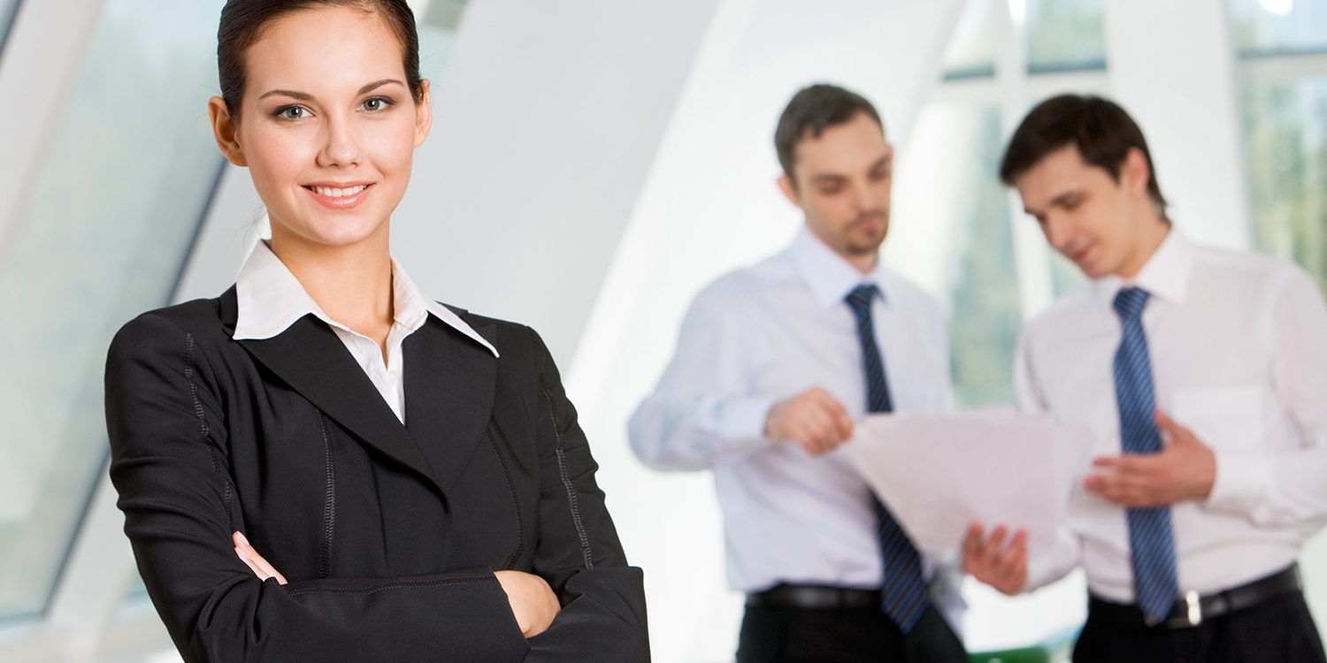 Professional businesswoman standing confidently with arms crossed and smiling, while two male colleagues review documents in the background inside a bright, modern office space.